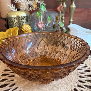 May include: A vintage amber-colored glass bowl with a diamond pattern and floral designs. The bowl has a scalloped rim and is likely used for serving or display. The bowl is sitting on a beige surface.
