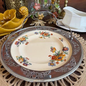 May include: A vintage serving plate with a floral design. The plate has a cream-colored center with colorful flowers and a silver-colored metal rim and handle. The flowers are in shades of orange, yellow, blue, and purple.