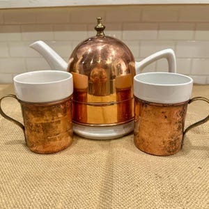 Puede incluir: Un juego de té de cobre con dos tazas y una tetera. La tetera tiene una cúpula de cobre y un asa y un pico blancos. Las tazas tienen interiores blancos y exteriores de cobre con asas de latón. El juego está sobre una superficie marrón.