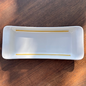 Könnte beinhalten: Eine rechteckige weiße Keramikschale mit abgerundeten Kanten. Das Design besteht aus zwei dünnen goldenen Streifen und einem dünnen marineblauen Streifen dazwischen, die entlang der oberen und unteren Kanten verlaufen. Die Schale ist leer und steht auf einer dunklen Holzoberfläche.