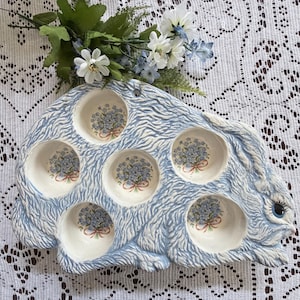 Könnte beinhalten: Ein Set aus drei Keramikbechern mit gesprenkelter, cremefarbener Glasur. Jeder Becher zeigt ein handgemaltes Blumendesign in Blau-, Grün- und Gelbtönen. Die Becher haben eine einfache, abgerundete Form und einen Griff.