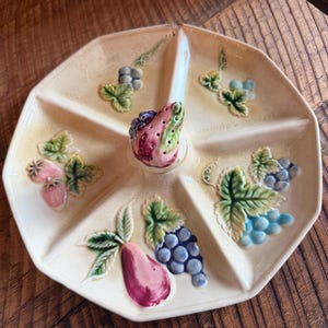 May include: A cream-coloured ceramic serving tray with five divided sections and a central handle. The tray features embossed, hand-painted fruit motifs including pink pears, green leaves, and clusters of blue and purple grapes in each section.