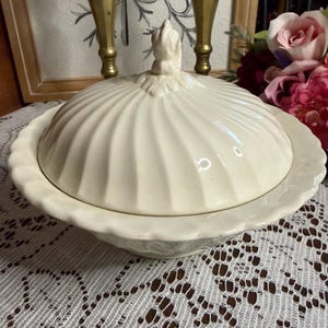 May include: Cream-colored, lidded serving dish with a scalloped edge. The lid features a ribbed design and a decorative finial. The dish is set on a white lace tablecloth, suggesting a vintage or antique aesthetic.