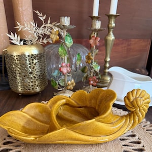 Könnte beinhalten: Eine vintage, senfgelbe Keramik-Servierschale mit blattartigem Design und Spiralgriff. Die Schale steht auf einer braunen Oberfläche, mit anderen dekorativen Gegenständen im Hintergrund, darunter Kerzenständer und eine Vase.