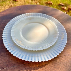 Puede incluir: Dos platos antiguos de vidrio opalino con borde festoneado y detalles florales dorados. Los platos están apilados, uno más pequeño sobre otro más grande. Los platos son de color blanco crema suave con un sutil tono azul.