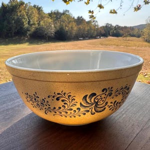 May include: A large, tan mixing bowl with a white interior and a black floral design around the exterior. The bowl is made of ceramic and sits on a wooden surface. The background shows a natural outdoor setting.