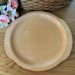 May include: A round, peach-colored serving platter with a white rim and decorative edge. The platter has two handles and is made of ceramic. The background includes flowers and a woven basket.