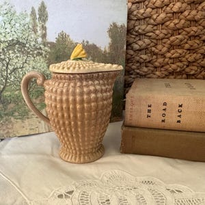 Puede incluir: Una cremera vintage de cerámica color beige con un patrón texturizado similar a burbujas. Tiene un asa curva y una tapa coronada por una flor amarilla. La cremera está sobre un paño blanco.