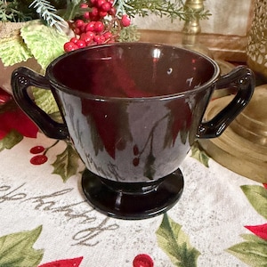 May include: A dark burgundy glass bowl with two handles, set on a pedestal base. The bowl has a glossy finish and reflects light. The bowl is likely vintage and suitable for serving. The background includes holiday decorations.