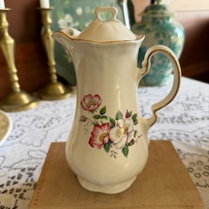 Op de afbeelding: Crèmekleurige keramische kan met een deksel, met een bloemenontwerp van roze en witte bloemen met groene bladeren. De kan heeft een gouden rand en een gebogen handvat. De kan staat op een houten oppervlak.