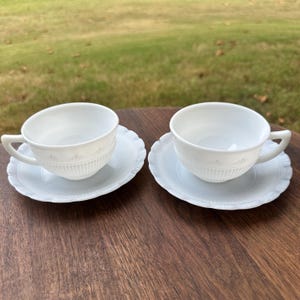 Puede incluir: Dos tazas de té y platillos blancos. Las tazas tienen un diseño acanalado en la mitad inferior y un asa curva. Los platillos tienen un borde festoneado y un patrón en relieve a juego. El juego está colocado sobre una superficie de madera.