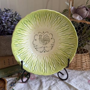 May include: A round ceramic bowl with a scalloped edge and a light green glazed finish. The center features a dark green line drawing of a rooster surrounded by a decorative circular border. The bowl has a textured, radiating leaf-like pattern.