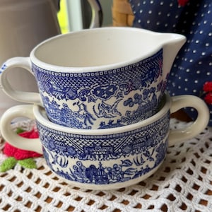 May include: Two stacked ceramic pieces featuring a blue and white willow pattern. The top piece is a creamer with a handle and spout, while the bottom piece is a bowl with two side handles. Both items display intricate landscape illustrations.