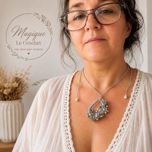 May include: A silver-toned crochet necklace featuring a teardrop-shaped pendant adorned with small teal crystals, white faux pearls, and intricate floral motifs. The necklace has a thin cord with two small white pearl drops at the ends.