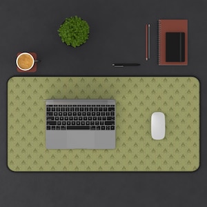May include: A large rectangular desk mat featuring a repeating olive green chevron pattern. The mat is positioned under a silver laptop and a white computer mouse. Surrounding items include a small potted plant, a coffee mug, a pen, and a notebook.