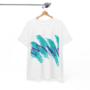 Könnte beinhalten: Ein weißes T-Shirt mit kurzen Ärmeln und einem zentralen Grafikdruck aus abstrakten, türkisfarbenen und violetten Pinselstrichen in einem diagonalen Muster. Das Shirt hängt auf einem Holzkleiderbügel an einer horizontalen Metallstange.