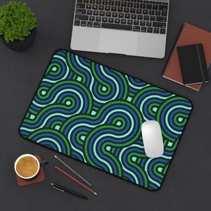 Op de afbeelding: Een rechthoekige muismat met een herhalend patroon van blauwe en groene gebogen lijnen op een zwarte achtergrond. Een witte computermuis ligt op de mat. Een laptop, notitieboekjes en een kopje koffie zijn ook zichtbaar.