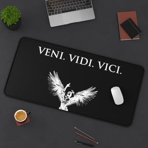 May include: A rectangular black desk mat featuring the white text "VENI. VIDI. VICI." centered at the top. Below the text is a white graphic illustration of a winged figure with arms raised, depicted in a sketch-like style against the black background.