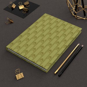 May include: A rectangular notebook with a muted olive green cover featuring a repeating pattern of darker green chevron arrows. The notebook is positioned alongside two pencils, one gold and one black, and several gold metal binder clips.