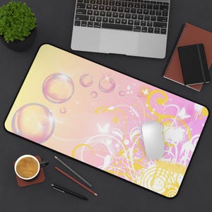 May include: A rectangular mousepad with a pink and yellow gradient background, featuring large bubble illustrations and white floral and butterfly designs. A white computer mouse rests on the pad, alongside a laptop, coffee cup, and stationery.