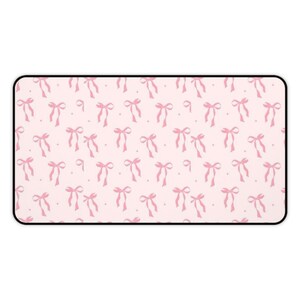 Puede incluir: Alfombrilla de ratón rectangular rosa con un patrón repetido de lazos rosas y pequeños puntos. Los lazos están atados y tienen cintas que fluyen. La alfombrilla tiene un borde negro.