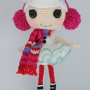 May include: A crocheted doll with white hair, a pink and white scarf, a black top hat with a pink ribbon, and pink pom-pom earmuffs. The doll is wearing a white dress with a blue trim and red and white striped leggings.