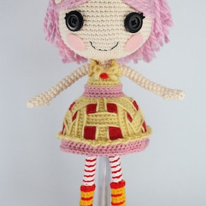 May include: A crocheted doll with pink hair, a yellow and red dress, and yellow and red striped socks. The doll is standing on a white stand.