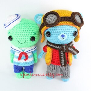 May include: Two crocheted stuffed toys, one green with a sailor hat and blue and white striped shirt, the other blue with a yellow aviator hat and brown goggles, wearing a red vest and gray pants.