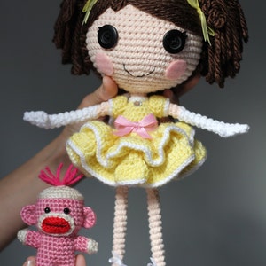 May include: A crocheted doll with brown hair, a yellow dress, and white shoes. The doll is holding a pink crocheted monkey.