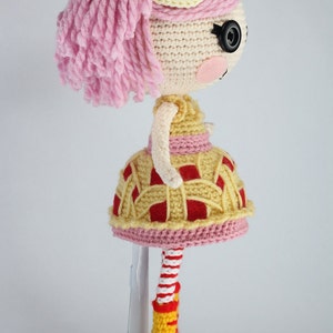 May include: A crocheted doll with pink hair, a yellow and red pie-shaped dress, and yellow boots. The doll is standing on a white stand.