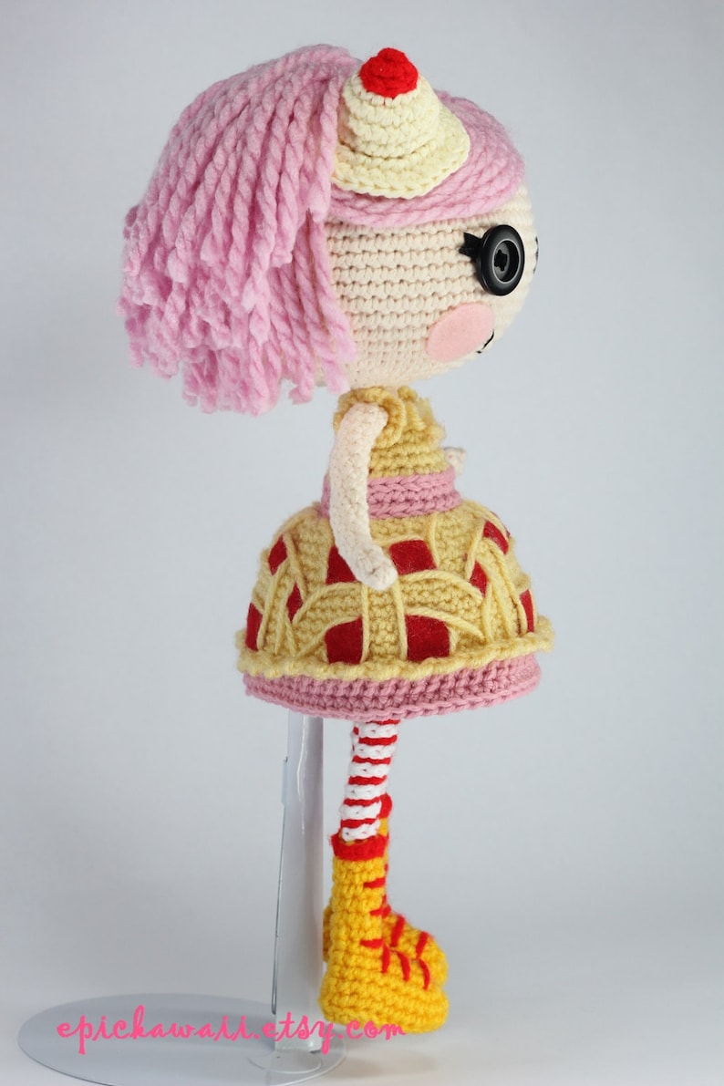May include: A crocheted doll with pink hair, a yellow and red pie-shaped dress, and yellow boots. The doll is standing on a white stand.