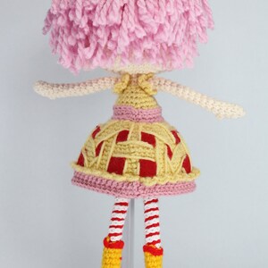 May include: A crocheted doll with pink hair, a yellow and red dress, and yellow boots. The doll is standing on a white stand.