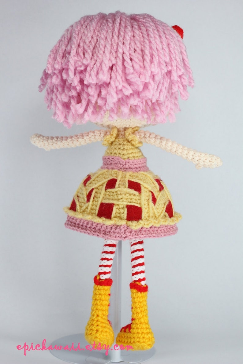 May include: A crocheted doll with pink hair, a yellow and red dress, and yellow boots. The doll is standing on a white stand.