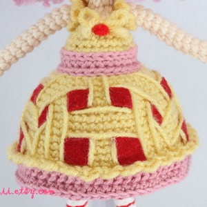 May include: Crocheted doll with pink hair wearing a yellow dress with red squares and a pink trim.