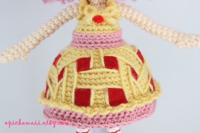 May include: Crocheted doll with pink hair wearing a yellow dress with red squares and a pink trim.