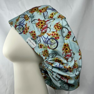 May include: A light blue surgical scrub hat with a repeating pattern of bicycles with baskets of sunflowers. The hat has a gathered back and a label that reads "Betty & Bella".