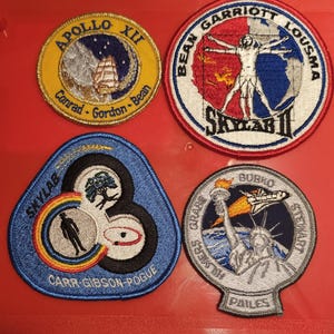 Peut inclure: Quatre écussons brodés de missions spatiales. En haut à gauche : Apollo XII avec un navire. En haut à droite : Skylab II avec une figure de l'Homme de Vitruve. En bas à gauche : Skylab 3 avec un arc-en-ciel. En bas à droite : Statue de la Liberté.