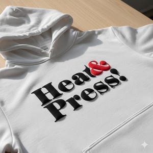 May include: A white hoodie featuring the text "Heat & Press!" in black, with a red ampersand. The sweatshirt has a front pocket and a drawstring hood. The garment is made of a soft material.