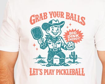 May include: A white short-sleeve t-shirt featuring a graphic of a cowboy holding a pickleball paddle and ball. The text reads GRAB YOUR BALLS, BIG DINK ENERGY!, and LET'S PLAY PICKLEBALL in red and blue ink with a desert cactus background.