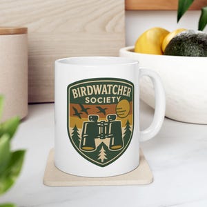 Könnte beinhalten: Weiße Keramiktasse mit einer grünen, braunen und gelben Grafik mit der Aufschrift "Birdwatcher Society". Das Design umfasst Silhouetten von Vögeln, Ferngläsern, einer Sonne und Bäumen. Die Tasse steht auf einem beigen Untersetzer.