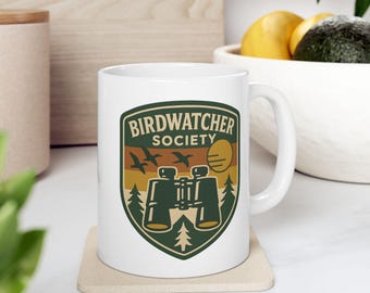 May include: White ceramic mug with a green, brown, and yellow graphic that reads "Birdwatcher Society." The design includes silhouettes of birds, binoculars, a sun, and trees. The mug sits on a beige coaster.