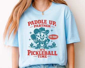 Può includere: T-shirt azzurra con grafica rossa e blu. La grafica presenta un personaggio di pickleball sorridente con cappello da cowboy che tiene una racchetta, con il testo "Paddle Up, Partner, It's Pickleball Time."