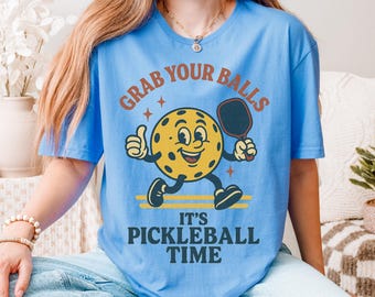 May include: Light blue t-shirt with the text "Grab Your Balls, It's Pickleball Time." The shirt features a cartoon pickleball character holding a paddle and giving a thumbs up. The design is in retro style.