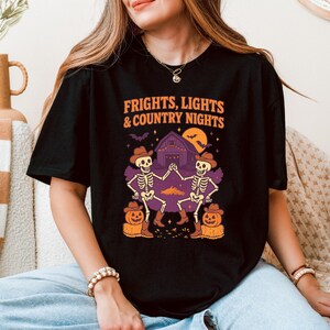 以下が含まれることがあります： オレンジ色の「FRIGHTS, LIGHTS & COUNTRY NIGHTS」の文字が入った黒いTシャツ。カウボーイハットをかぶった2体の骸骨が、納屋、カボチャ、満月の前で踊っているグラフィック。楽しいハロウィーンをテーマにしたデザイン。