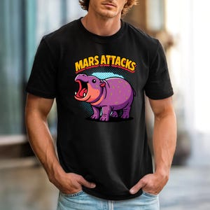 以下が含まれることがあります： 漫画のイラストが描かれた黒いTシャツ。紫色のカバが描かれており、ピンクとオレンジのアクセントが施されています。「MARS ATTACKS」の文字が黄色と赤色でカバの上に表示されています。