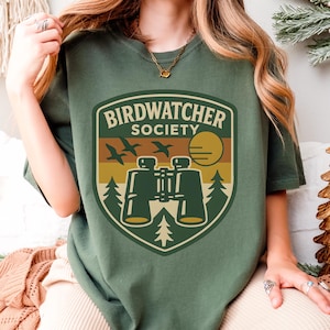 May include: Green t-shirt with a "Birdwatcher Society" graphic. The design features binoculars, birds, a sun, and trees in a shield shape. The text is in a vintage style, perfect for nature enthusiasts.
