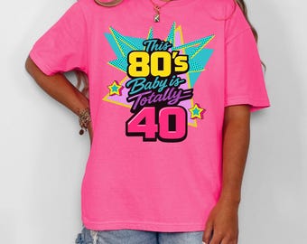 May include: A bright pink t-shirt with the text "This 80's Baby is Totally 40" in a colorful retro design. The graphic includes stars and a burst of teal and yellow shapes. The shirt is a casual, oversized fit.