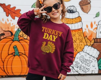 Op de afbeelding: Een kind draagt een bordeauxrode sweatshirt met de tekst "TURKEY DAY" en een kalkoenafbeelding. Het kind draagt een zonnebril en een zwarte korte broek, en maakt een vredesteken. De achtergrond toont een muurschildering met pompoenen en herfstbladeren.