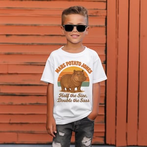 May include: White t-shirt featuring a cartoon hippo with the text "Mars Potato Jones" above and "Half the Size, Double the Sass" below. The hippo is brown, set against a sunset design. The shirt is worn by a child.