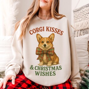 May include: A beige sweatshirt featuring a corgi illustration wearing a plaid bow tie. The text "CORGI KISSES" is arched above the corgi, and "& CHRISTMAS WISHES" is below. The sweatshirt is part of a holiday-themed collection.